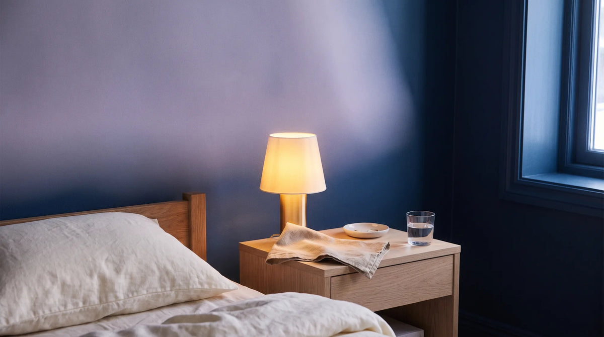 Chevet au crépuscule : lampe allumée, verre d'eau, linge plié et coin de lit en lin crème